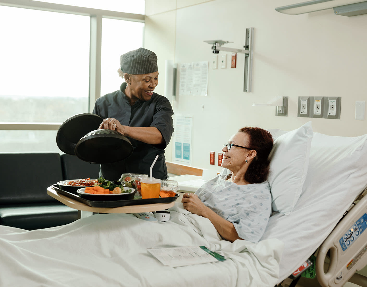 A nurse delivers a food tray to a woman in a hospital bed.